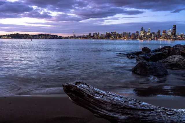 A driftwood log on dark sand in blue twilight, with the lit downtown skyline across Elliott Bay under purple clouds.
