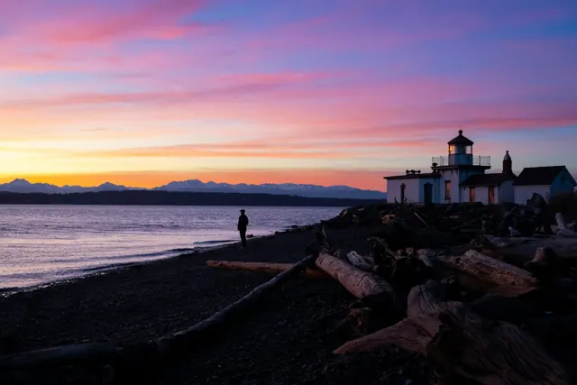 A pink and orange sunset over calm water and distant mountains, with the small white West Point Lighthouse and driftwood on the beach.