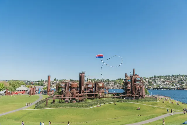 Rust-red towers and pipes on a grassy hill, people on the lawns, a kite in a clear sky, and Lake Union behind.